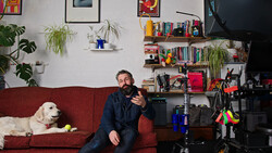 Man sitting on red couch with white dog, gesturing while speaking in a home studio setup.