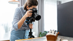 Female photographer holding a mirrorless camera with telephoto lens mounted on a tripod, shooting a styled food setup.