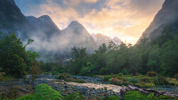 Mountain valley at sunrise with river, mist, and dense forest beneath towering peaks.