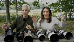 Two people display a collection of camera lenses arranged on a wooden table outdoors.