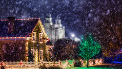 Snow falling heavily over a illuminated gingerbread house with a white picket fence and a historic cathedral in the background at night.