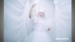Woman in white ethereal dress holding ornate birdcage amid flowing white fabric and soft lighting.