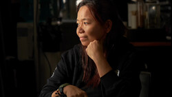 Woman in dark jacket with hand near face, contemplative expression in dim interior setting.