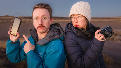 Two photographers in winter clothing posing outdoors with a smartphone and compact camera against a barren landscape.