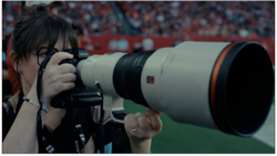 Sports photographer with telephoto lens at a stadium event with crowd in background.