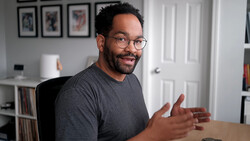 Man wearing glasses and dark t-shirt gesturing while speaking in a home office setting.