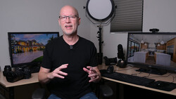 Bald man with glasses speaking to camera in a photography studio setup with dual monitors and professional lighting equipment.