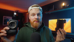 Photographer holding a camera body in one hand and a smartphone in the other, displayed in a studio with warm and cool ambient lighting.