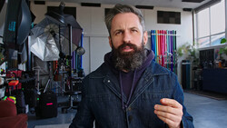 Bearded man in denim jacket inside a photography studio with lighting equipment and reflectors visible behind him.