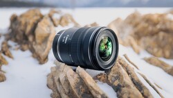 Wide-angle zoom lens positioned on rocky terrain with snowy background and blurred mountains.