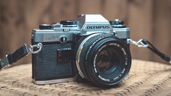 Olympus OM10 35mm film camera with black leather grip and attached Zuiko lens on woven surface.