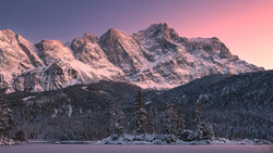 Snow-covered alpine peaks glowing pink at sunset above a frozen lake and evergreen forest.