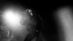 Singer performing on stage with microphone, backlit by intense stage lighting against dark background.