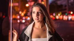 Woman in dark jacket leaning against neon sign with blurred city lights in background.
