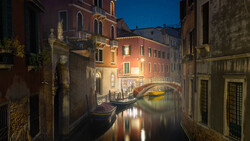 Venetian canal at twilight with illuminated palazzo buildings, gondola, and arched bridge reflecting in calm water.