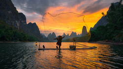 Fisherman casting net at sunrise among dramatic limestone karst formations reflected in still water.