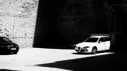 White sedan parked in stark sunlight against concrete wall with deep shadow in monochrome.