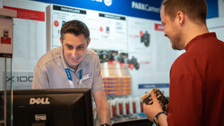 Two men at a tech conference booth discussing equipment, one wearing a lanyard badge at a Dell computer workstation.