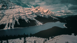 Snow-covered mountain peaks tower above a turquoise alpine lake surrounded by evergreen forest.