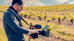 Videographer operating a professional camera rig with microphone and monitor in a vineyard during golden hour.