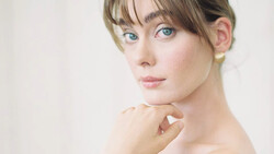 Woman with blue eyes and bangs gazing toward camera with hand near face.