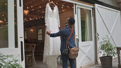 Woman in dark blazer examining a white lace wedding dress hanging indoors.
