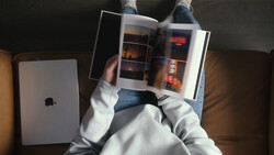 Overhead view of a person in a white hoodie holding an open photography magazine or portfolio while sitting on a couch with an iPad nearby.