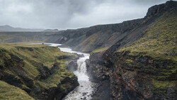 Glacial river cutting through moss-covered canyon with basalt cliffs under overcast sky.