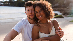 Couple embracing on a sandy beach during golden hour with water and shoreline in background.