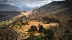 Rolling mountains and valleys with scattered trees and winding paths under dramatic cloudy sky.