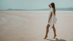 Woman in white dress standing barefoot on expansive sandy beach, gazing toward distant horizon and mountains.