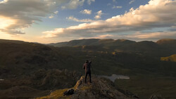 Photographer on rocky outcrop with tripod overlooking mountainous valley at golden hour.