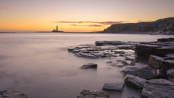 Rocky coastline at sunset with calm water and lighthouse in the distance.