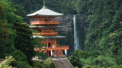 Red-and-gold pagoda with multiple tiers nestled against a tall waterfall in a forested canyon.
