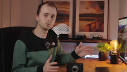 Man in green and black shirt gesturing while speaking at desk with camera and laptop.