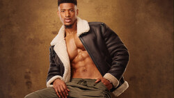 Shirtless man in black leather jacket with sherpa collar posing against textured brown backdrop.