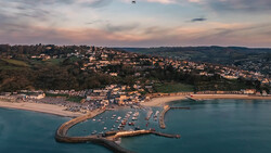 Coastal town with curved harbor pier at golden hour, surrounded by hillside homes and sandy beach.
