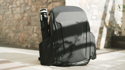 Black hard-shell camera backpack photographed outdoors with natural sunlight creating strong shadows on pavement.