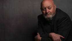 Bald man with beard in dark clothing against neutral gray background.