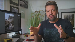 Man gesturing while discussing photography at a desk with a monitor displaying landscape imagery.