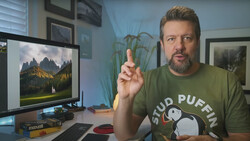 Man in green shirt gesturing while speaking at a desk with a monitor displaying mountain landscape photography.