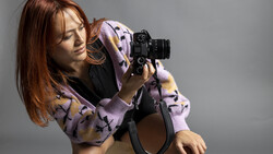 Woman holding a film camera to her eye, wearing a lavender patterned cardigan against a neutral gray background.