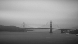 Suspension bridge spanning a body of water under overcast skies in black and white.