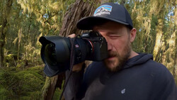 Photographer holding a DSLR camera with a telephoto lens in a mossy forest setting.