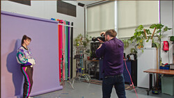 Cinematographer filming a portrait subject against a purple backdrop in a studio with professional lighting equipment.