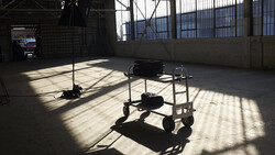 Film production equipment including lighting rigs and camera carts arranged on a concrete stage floor in an industrial studio space.