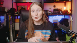 Woman with reddish-brown hair wearing a black graphic tee, seated at a desk in a gaming setup with colorful ambient lighting.