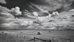 Expansive prairie landscape with wooden ranch fence in foreground and dramatic cloudy sky overhead.
