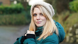 Woman in teal jacket and cream beanie smiling at camera outdoors near water.