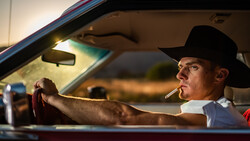 Man in cowboy hat smoking cigarette while driving vintage car at sunset.